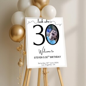 Peut inclure: Un panneau blanc sur un chevalet en bois avec le texte "look who's 30 Welcome to STEVEN S 30TH BIRTHDAY" et une photo d'un enfant. Le panneau est d&eacute;cor&eacute; de ballons dor&eacute;s et blancs.