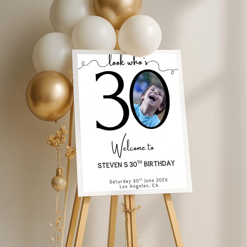 Peut inclure: Un panneau blanc sur un chevalet en bois avec le texte "look who's 30 Welcome to STEVEN S 30TH BIRTHDAY" et une photo d'un enfant. Le panneau est d&eacute;cor&eacute; de ballons dor&eacute;s et blancs.