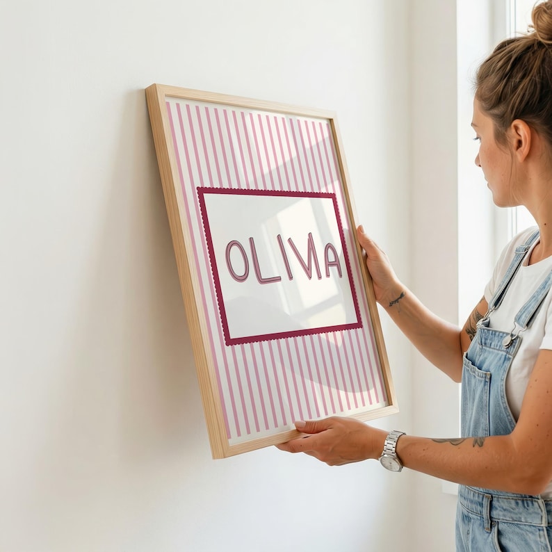 May include: A rectangular framed wall art piece featuring a light pink and white vertical striped background. Centered on the print is a white rectangular panel with a scalloped dark pink border containing the name OLIVA in dark pink letters.