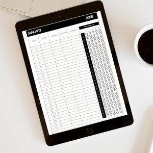Op de afbeelding: Een zwarte tablet met een planner voor januari 2026 met kolommen voor dagelijkse invoer en een stemmingbeoordelingssectie. Een kopje koffie en een pen liggen naast de tablet op een wit oppervlak.
