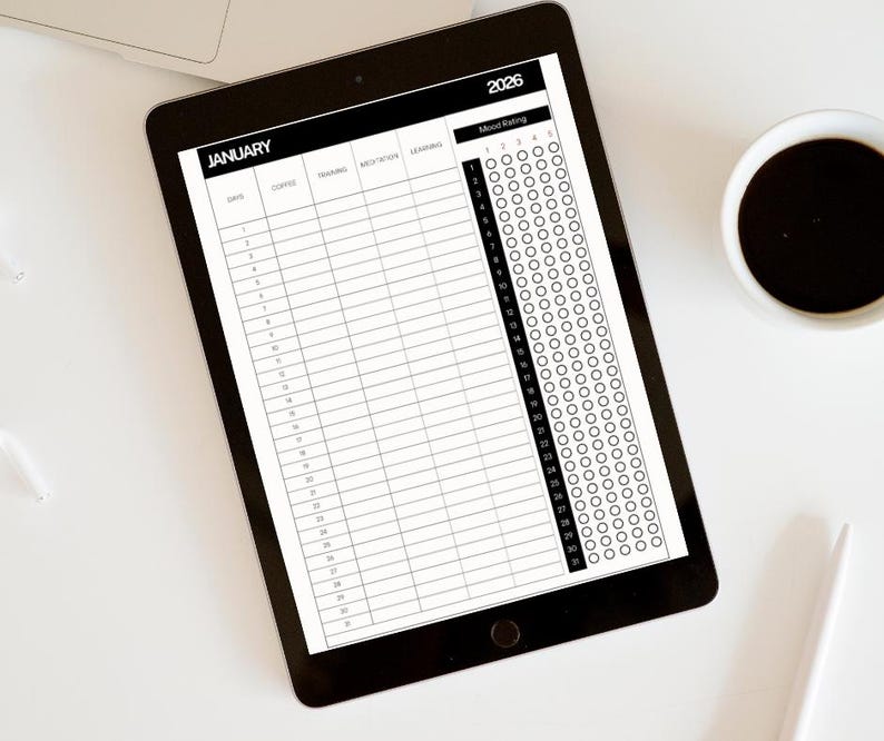 Op de afbeelding: Een zwarte tablet met een planner voor januari 2026 met kolommen voor dagelijkse invoer en een stemmingbeoordelingssectie. Een kopje koffie en een pen liggen naast de tablet op een wit oppervlak.
