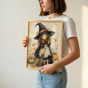 Puede incluir: Una l&aacute;mina art&iacute;stica enmarcada con una ilustraci&oacute;n en acuarela de un personaje joven con un sombrero de bruja negro puntiagudo, un vestido oscuro con delantal blanco y una escoba. Un peque&ntilde;o gato negro se sienta cerca entre calabazas naranjas.