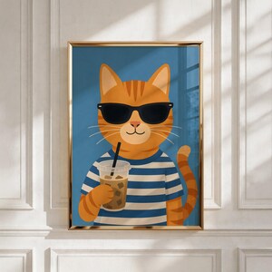 May include: A framed wall art print featuring an orange tabby cat wearing black sunglasses and a blue and white striped shirt. The cat is holding a clear plastic cup filled with an iced beverage and a black straw against a solid blue background.