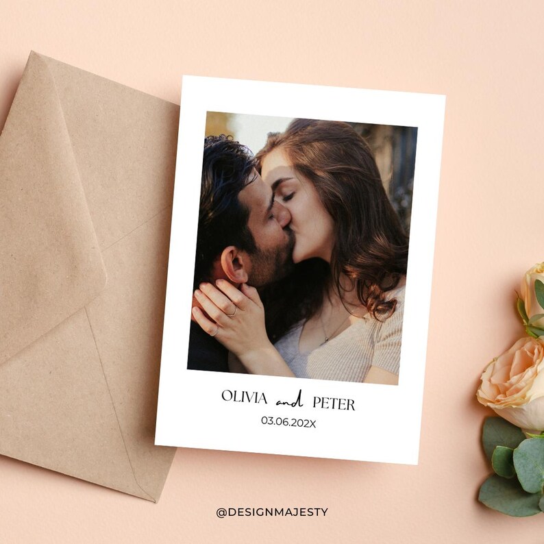 Op de afbeelding: Een witte kaart met een foto van een kussend stel, met de namen "OLIVIA and PETER" en de datum "03.06.202X" eronder gedrukt. Een bruine envelop staat links en bloemen rechts. De kaart heeft de tekst "@DESIGNMAJESTY" onderaan.