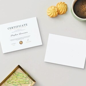 以下が含まれることがあります： 白い参加証明書に「CERTIFICATE OF PARTICIPATION」と「Stephen Lawrence」というテキストが黒で印刷されています。証明書は、コーヒーカップ、クッキー2つ、緑色のクリップが置かれた金のトレイと一緒に白い表面に置かれています。