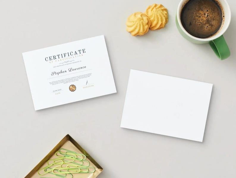 以下が含まれることがあります： 白い参加証明書に「CERTIFICATE OF PARTICIPATION」と「Stephen Lawrence」というテキストが黒で印刷されています。証明書は、コーヒーカップ、クッキー2つ、緑色のクリップが置かれた金のトレイと一緒に白い表面に置かれています。