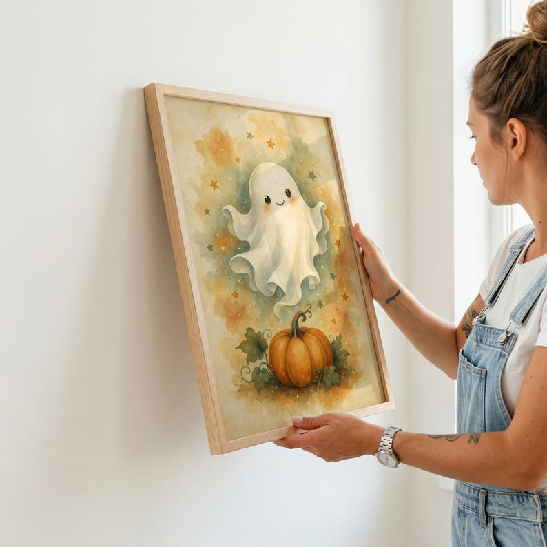 May include: A framed wall art print featuring a watercolor illustration of a smiling white ghost floating above a bright orange pumpkin. The background is a soft, mottled yellow and gold wash decorated with small, scattered brown and gold stars.