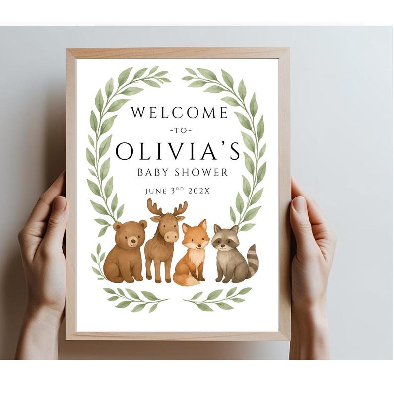 May include: A white sign with a green leafy wreath around the edges. The sign says "Welcome to Olivia's Baby Shower June 3rd 202X". There are four woodland animals in the center of the wreath: a bear, a moose, a fox, and a raccoon.