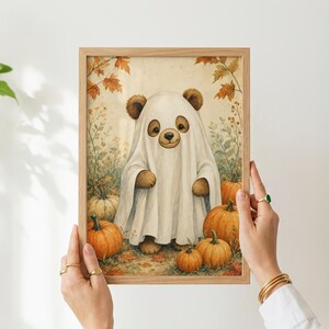 May include: A framed wall art print featuring a brown bear wearing a white ghost costume, standing in a field of orange pumpkins and autumn leaves. The illustration uses soft, warm tones to depict the bear and the surrounding fall harvest scene.