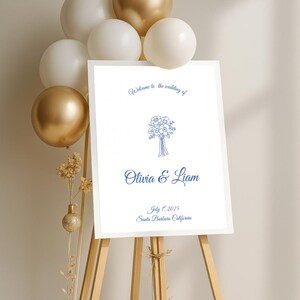May include: A white wedding welcome sign on a wooden easel, with blue text that reads "Welcome to the wedding of Olivia & Liam, July 1st 2025, Santa Barbara California." Gold and white balloons are in the background.