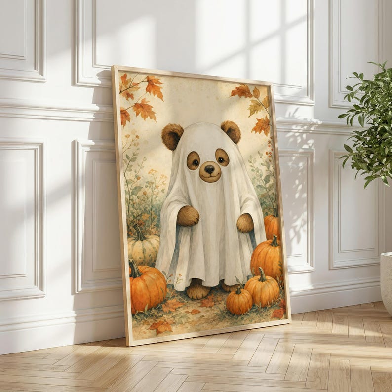 May include: A framed wall art print featuring a brown bear wearing a white ghost costume, standing in a field of orange pumpkins and autumn leaves. The illustration has a soft, warm color palette with golden tones and detailed fall foliage.