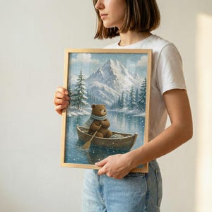 May include: A framed rectangular wall art print featuring a brown bear wearing a blue and white striped scarf, sitting in a wooden rowboat on a calm lake. The background displays snow-covered mountains and pine trees during a light snowfall.