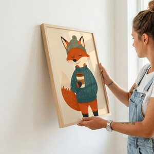 May include: A framed wall art print featuring a cartoon fox with orange fur, wearing a dark green knit beanie and a matching dark green sweater, holding a brown coffee cup. The illustration is set against a light cream background.