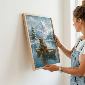 May include: A framed wall art print featuring a brown bear wearing a blue scarf, rowing a wooden boat on a calm lake. The background displays snow-covered mountains and pine trees under a light snowfall, captured in a vertical wooden frame.