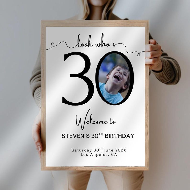 Peut inclure: Panneau encadr&eacute; avec le texte "look who's 30" et "Welcome to STEVEN S 30TH BIRTHDAY". Le chiffre 30 est grand et noir, avec une photo d'un enfant riant dans le z&eacute;ro. La date et le lieu sont &eacute;galement inclus.