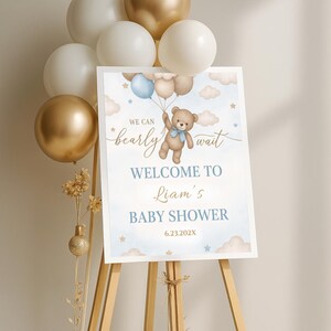 May include: A blue and white baby shower welcome sign with a teddy bear holding balloons. The sign says "We can bearly wait" and "Welcome to Liam's Baby Shower 6.23.202X".