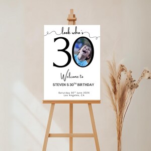 Peut inclure: Un panneau blanc sur un chevalet en bois avec le texte "look who's 30" et "Welcome to STEVEN S 30TH BIRTHDAY". Une photo d'une personne est dans le chiffre 0. La date et le lieu sont &eacute;galement imprim&eacute;s sur le panneau.