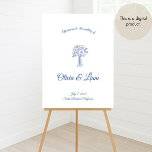 May include: A white wedding welcome sign on a wooden easel. The sign features blue script that reads "Welcome to the wedding of Olivia & Liam" with the date July 1st, 2025, and Santa Barbara, California. A digital product.
