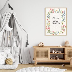May include: A floral-themed bridal shower sign with the text "Welcome to Olivia's Bridal Shower 13.05.2025". The sign is framed in a light wood frame and is hanging on a white wall.