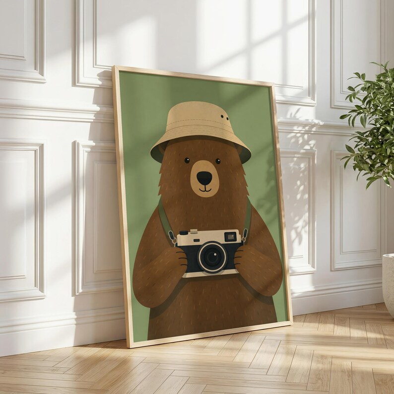 May include: A framed wall art print featuring a brown bear wearing a tan bucket hat and holding a vintage-style camera with a black and white body. The bear is centered against a solid olive green background within a light wood frame.