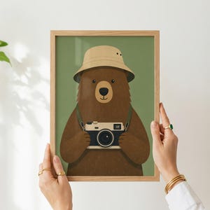 May include: A framed wall art print featuring a brown bear wearing a beige bucket hat and holding a vintage-style camera around its neck. The bear is centered against a solid olive green background, enclosed in a light-colored wooden frame.