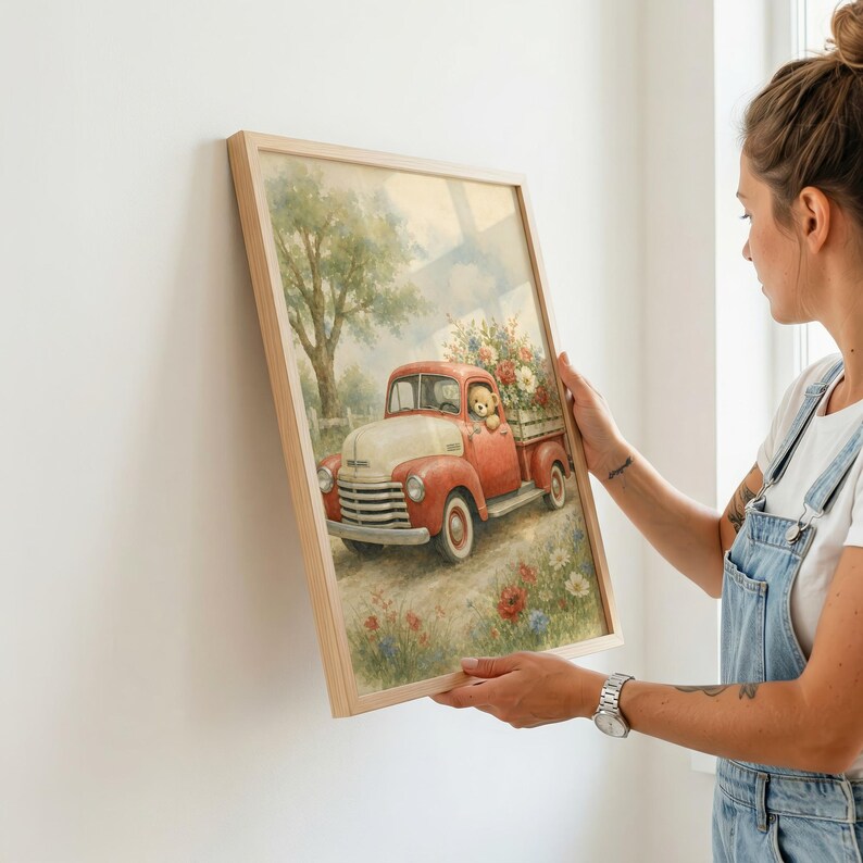 May include: A framed wall art print featuring a vintage red pickup truck with a cream-colored hood, carrying a load of colorful flowers in the bed. A teddy bear sits in the driver's seat. The scene is set in a field with trees and wildflowers.