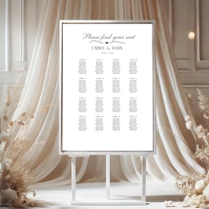 K&ouml;nnte beinhalten: Wei&szlig;e Hochzeitssitzordnung mit silbernem Rahmen auf einem wei&szlig;en Staffelei. Die Karte hat den Text "Bitte finden Sie Ihren Platz" und "Emma & John" mit einem kleinen Herzsymbol. Die Karte listet Tischnummern und G&auml;stelisten auf.