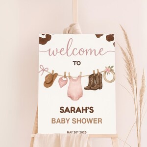 Może przedstawiać: Tablica powitalna na baby shower Sarah z brązowym tłem w kratę krowią. Na tablicy znajduje się r&oacute;żowy śpioszek dla niemowlęcia, brązowy kowbojski kapelusz i para brązowych kowbojskich but&oacute;w wiszących na sznurku do suszenia ubrań z r&oacute;żowymi kwiatami. Data baby shower to 20 maja 2025.