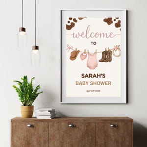 Może przedstawiać: Drukowana tablica powitalna na baby shower z brązowym tłem w kratę krowią. Tablica przedstawia sznurek do suszenia z r&oacute;żowym body dla dziecka, brązowym kowbojskim kapeluszem, r&oacute;żowym śliniakiem, brązowymi kowbojskimi butami i brązowym sznurkiem z r&oacute;żową kokardką. Tekst na tablicy brzmi "Witamy na baby shower Sarah 20 maja 2025".