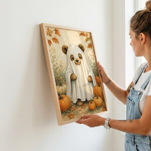 May include: A framed wall art print featuring a brown bear wearing a white ghost costume, standing in a field with orange pumpkins and autumn leaves. The illustration has a soft, light-colored background with fall foliage and scattered pumpkins.