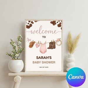 Może przedstawiać: Witamy na Baby Shower Sarah z r&oacute;żowo-brązowym tłem w krowie cętki. Na plakacie znajduje się sznurek do suszenia z kowbojskim kapeluszem, śliniakiem, r&oacute;żową dziecięcą koszulką i dwiema parami brązowych kowbojskich but&oacute;w.