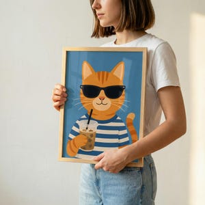 May include: A framed art print featuring a cartoon orange tabby cat wearing black sunglasses and a blue and white striped shirt. The cat is holding a clear plastic cup filled with an iced beverage and a black straw against a solid blue background.