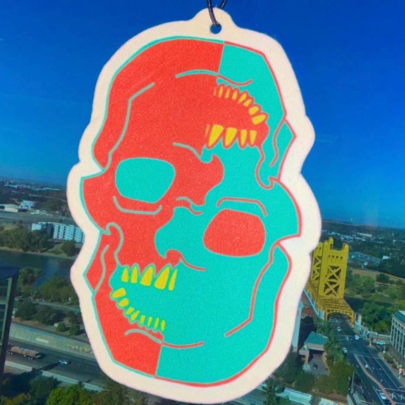 May include: A white car air freshener shaped like a skull. The skull is split in half, with one side red and the other side turquoise. The teeth are yellow.