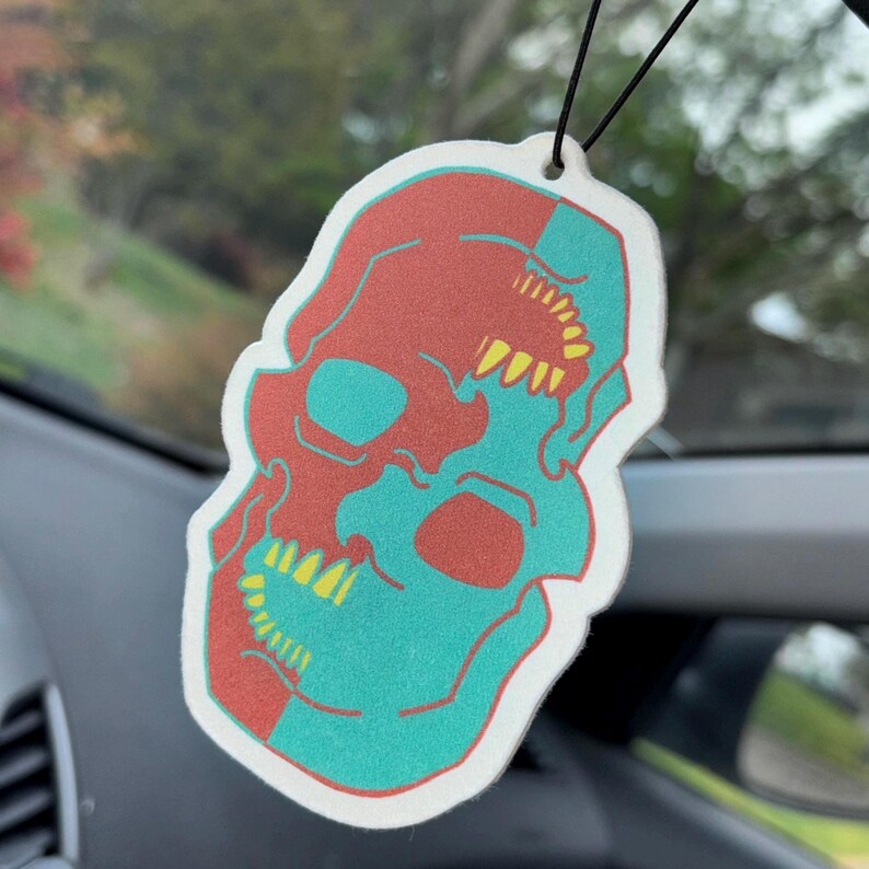 May include: A white car air freshener shaped like a skull with a red and teal colour scheme. The skull has yellow teeth and is split down the middle.