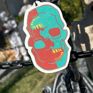 May include: A red and teal skull car air freshener with yellow teeth. The skull is split down the middle, with one side red and the other teal.