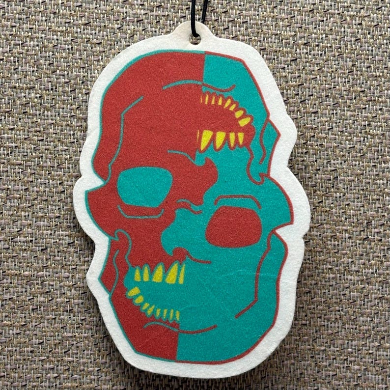 May include: A white car air freshener featuring a skull design. The skull is split in half, one side red and the other teal. The teeth are yellow.