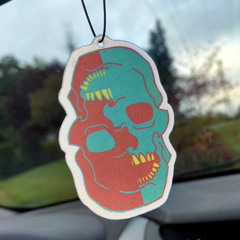 May include: A white car air freshener shaped like a skull with a red and teal colour scheme. The skull has yellow teeth and is split down the middle.