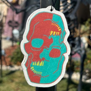 May include: A white car air freshener shaped like a skull with a red and teal colour scheme. The skull has yellow teeth and eye sockets.