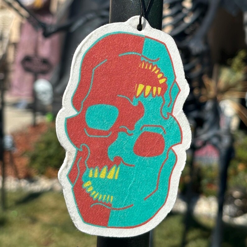 May include: A white car air freshener shaped like a skull with a red and teal colour scheme. The skull has yellow teeth and eye sockets.