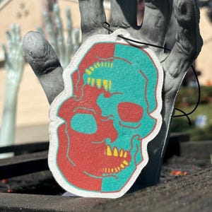 May include: A white car air freshener shaped like a skull with a red and teal colour scheme. The skull is split down the middle, with one side red and the other teal.