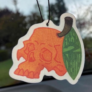 May include: Orange and green car air freshener shaped like a skull with a leaf. The skull has a large eye socket and a small mouth. The leaf is green with a brown stem.