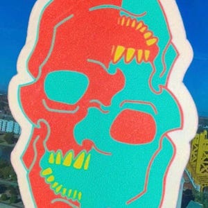 May include: A white car air freshener shaped like a skull. The skull is split in half, with one side red and the other side turquoise. The teeth are yellow.