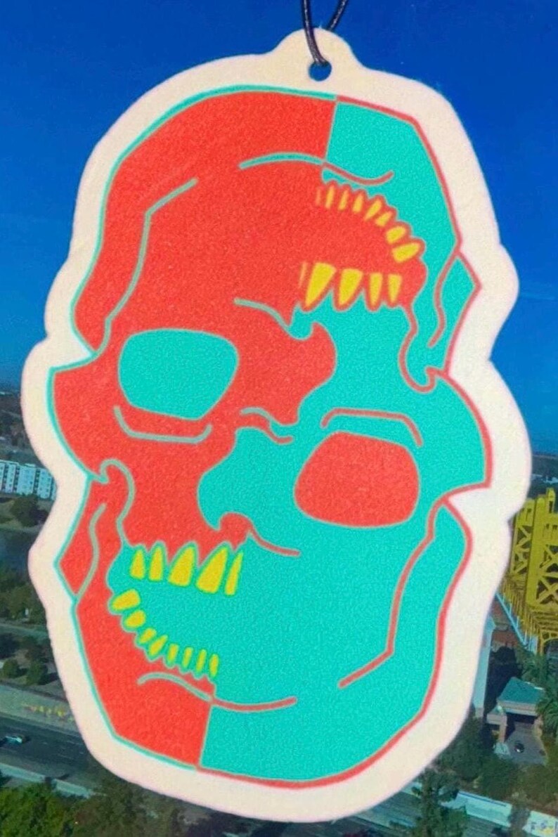 May include: A white car air freshener shaped like a skull. The skull is split in half, with one side red and the other side turquoise. The teeth are yellow.