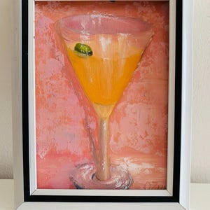 May include: An oil painting in a white and black frame depicting a martini glass filled with a vibrant orange liquid and a single green olive. The background is a textured, mottled pink and peach surface with visible brushstrokes throughout.