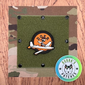 May include: A green patch with a cartoon pilot in a plane with the text "DOOFER" on a circle. The patch is attached to a green velcro panel with black velcro dots. A white circle with the text "Bubba's Patches" and a cartoon dog wearing aviator sunglasses is in the lower right corner.