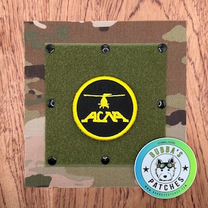 May include: A yellow and black embroidered patch with a helicopter and the text "TH-78 ACNA". The patch is attached to a green fabric background with black velcro dots.