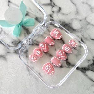 May include: A set of pink press-on nails with white floral designs, displayed in a clear plastic case. The nails have a matte finish and are oval-shaped. A light blue ribbon and a clear, round object are attached to the case.