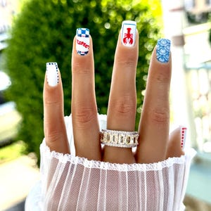 May include: A hand with manicured nails featuring various designs. One nail has a red lobster, another has a blue and white checkered pattern with text, and another has a blue seashell design. A gold ring with yellow stones is also visible.