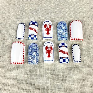 May include: A collection of ten white ceramic nail art designs. The designs include red lobsters, blue and white seashell patterns, and the word "Lobster" in red and blue checkered patterns. Some nails have red or blue dot borders.