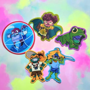 May include: A collection of five colorful vinyl stickers featuring cartoon characters. One is a circular blue turtle in a basketball hoop, another a character in a green cape, a frog-hooded figure, a turtle with a camera, and a blonde turtle with a sword.